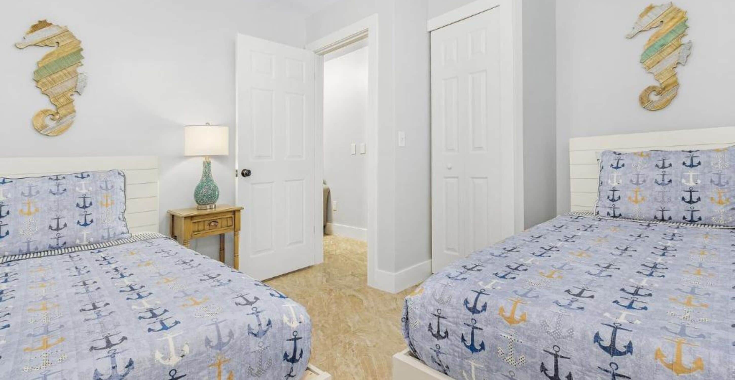 Un dormitorio con dos camas, cada una con una colcha azul con motivos de anclas, paredes blancas y decoración de caballitos de mar en la pared.