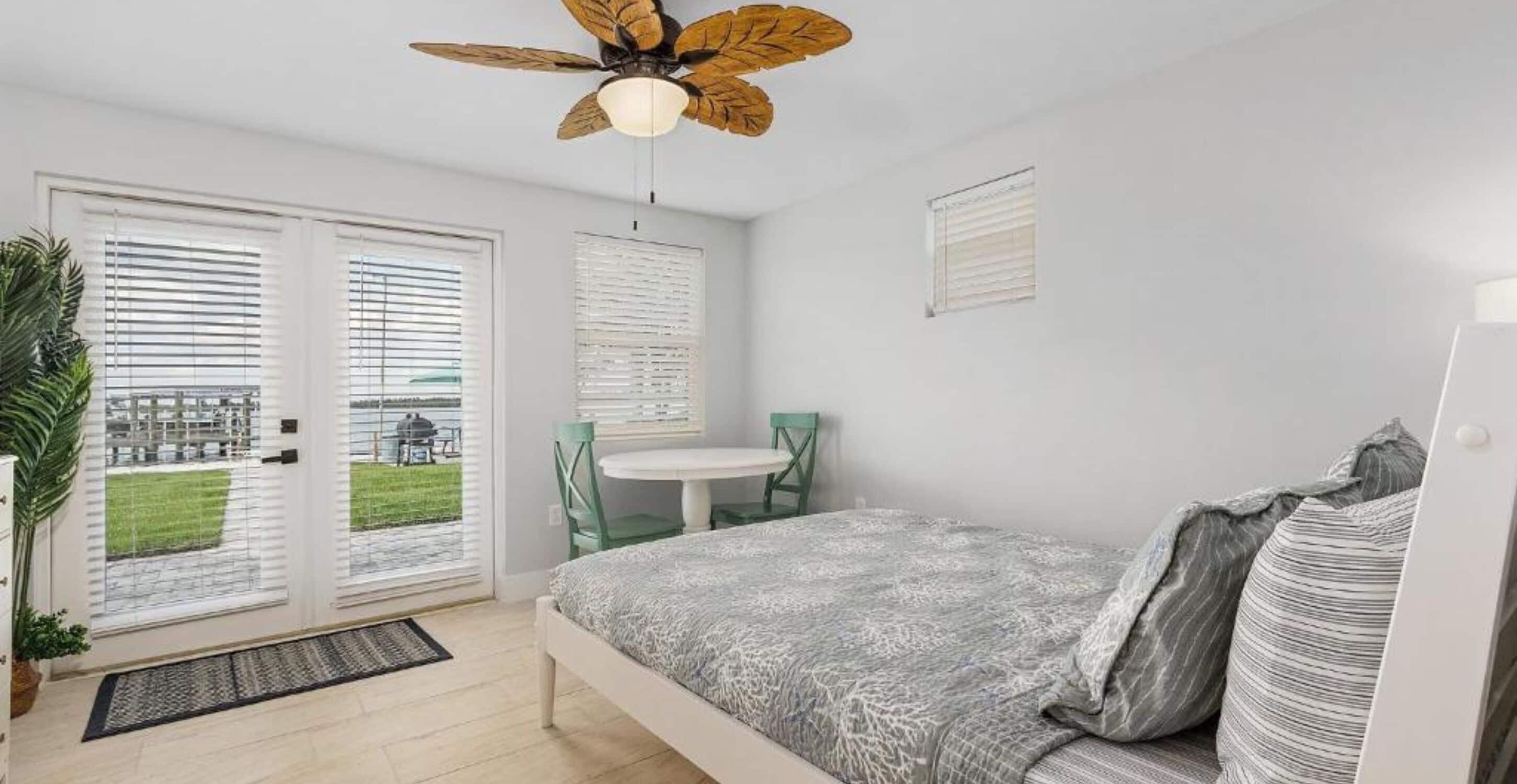 A bedroom with a bed, a ceiling fan, a window with blinds, and a view of the outside.