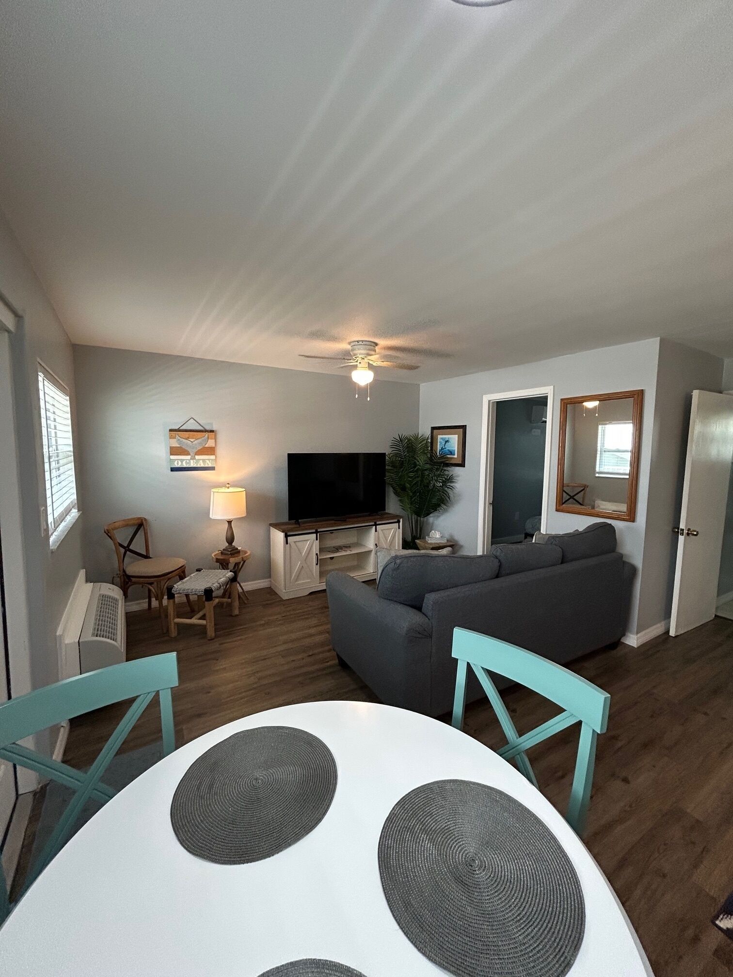 Un salon avec un canapé gris, un téléviseur, un ventilateur de plafond et un coin repas comprenant une table blanche et des chaises turquoise.