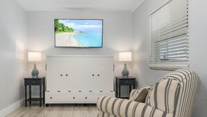 A bedroom with a large bed, a TV mounted on the wall, two bedside tables with lamps, and a window with blinds. - Matlacha on Shoreview (Cape Coral)