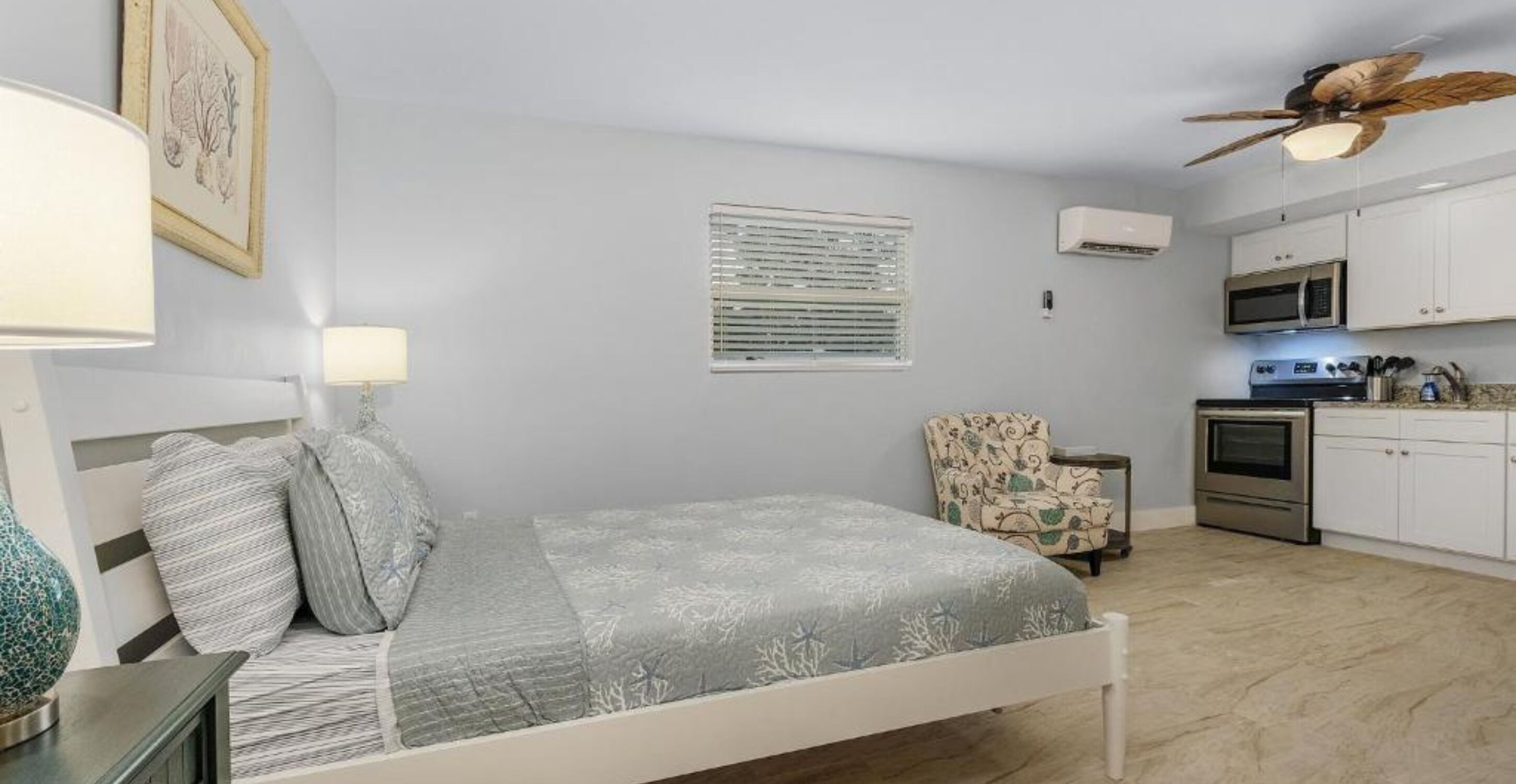A bedroom with a bed, bedside lamps, a chair, a window with blinds, an air conditioning unit, a ceiling fan, and a kitchenette with a microwave and stove.