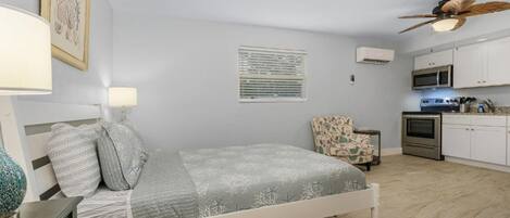 A bedroom with a bed, bedside lamps, a chair, a window with blinds, an air conditioning unit, a ceiling fan, and a kitchenette with a microwave and stove.