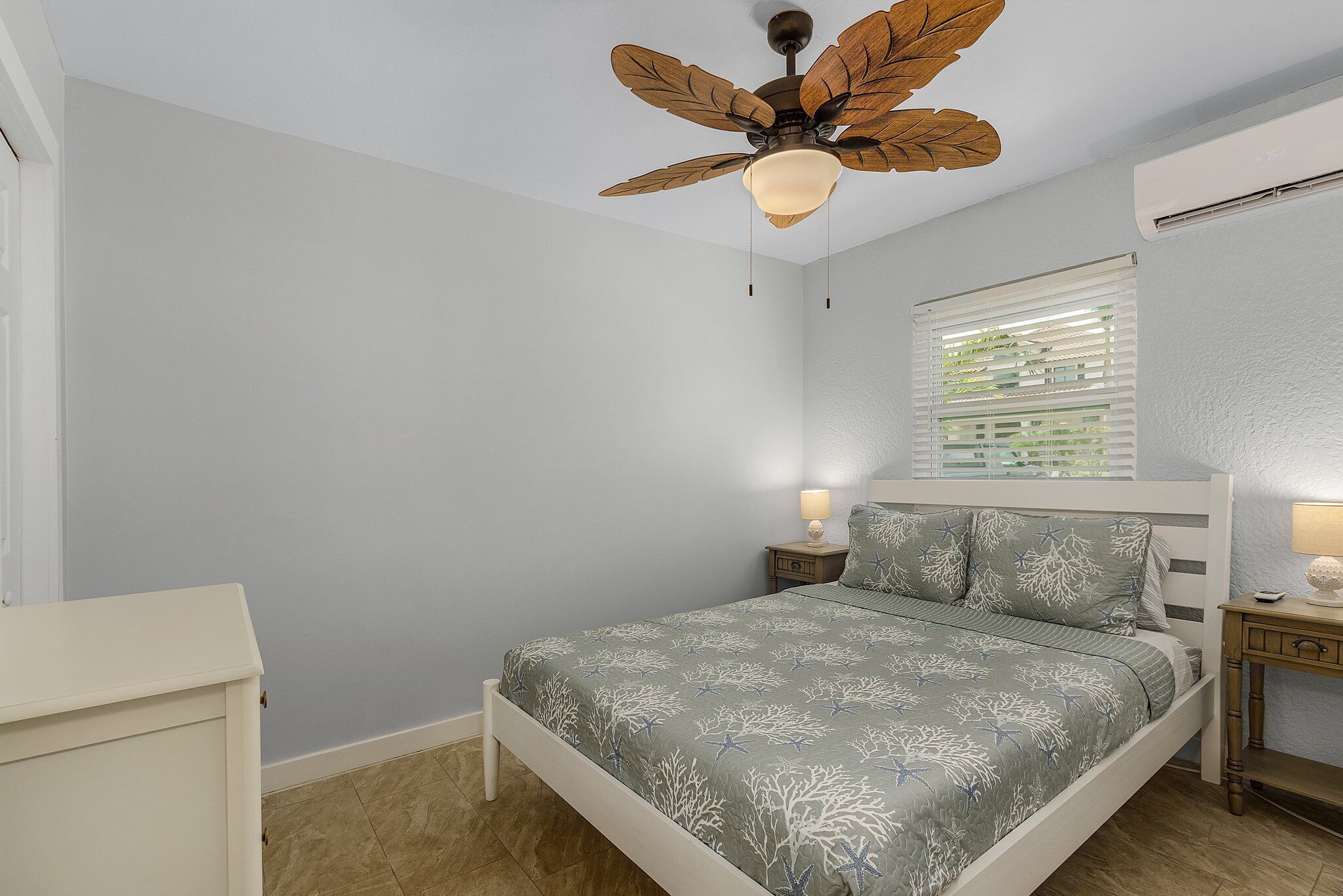 Une chambre à coucher comprenant un lit, un ventilateur de plafond, une fenêtre avec des stores, une table de chevet avec une lampe et une commode.