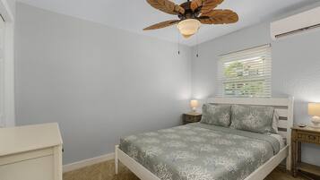 Une chambre à coucher comprenant un lit, un ventilateur de plafond, une fenêtre avec des stores, une table de chevet avec une lampe et une commode.