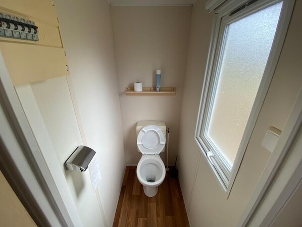 A small, narrow bathroom with a toilet, a shelf with toiletries, and a window. - Mobile Homes at Camping Parc des Roches (Saint-Chéron)
