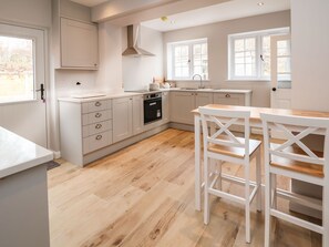 A modern kitchen with white cabinetry, a built-in oven, and a dining area with white chairs and a table. - 2 Golf Links Cottages (Northwich)