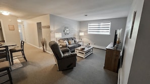 A living room with a sofa, armchair, coffee table, and a TV stand. There is a dining area with chairs and a table.