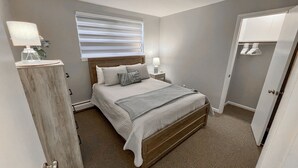 Un dormitorio con cama, lámparas de noche, ventana con persianas, cómoda de madera y una puerta.