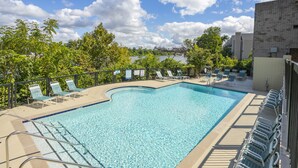 A rectangular swimming pool with clear blue water, surrounded by a black metal fence. There are multiple lounge chairs along the poolside, and the pool area is adjacent to a building with a tiled exterior. - ❤️ of DWNTWN, Riverfront w pool, walk 2 Broadway (Nashville)