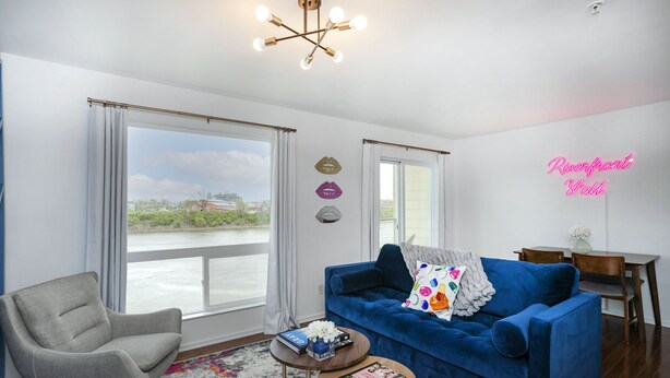 A modern living room with a blue sofa, a grey armchair, and a coffee table. There is a large window with curtains, a neon sign, and a view of a body of water. - ❤️ of DWNTWN, Riverfront w pool, walk 2 Broadway (Nashville)