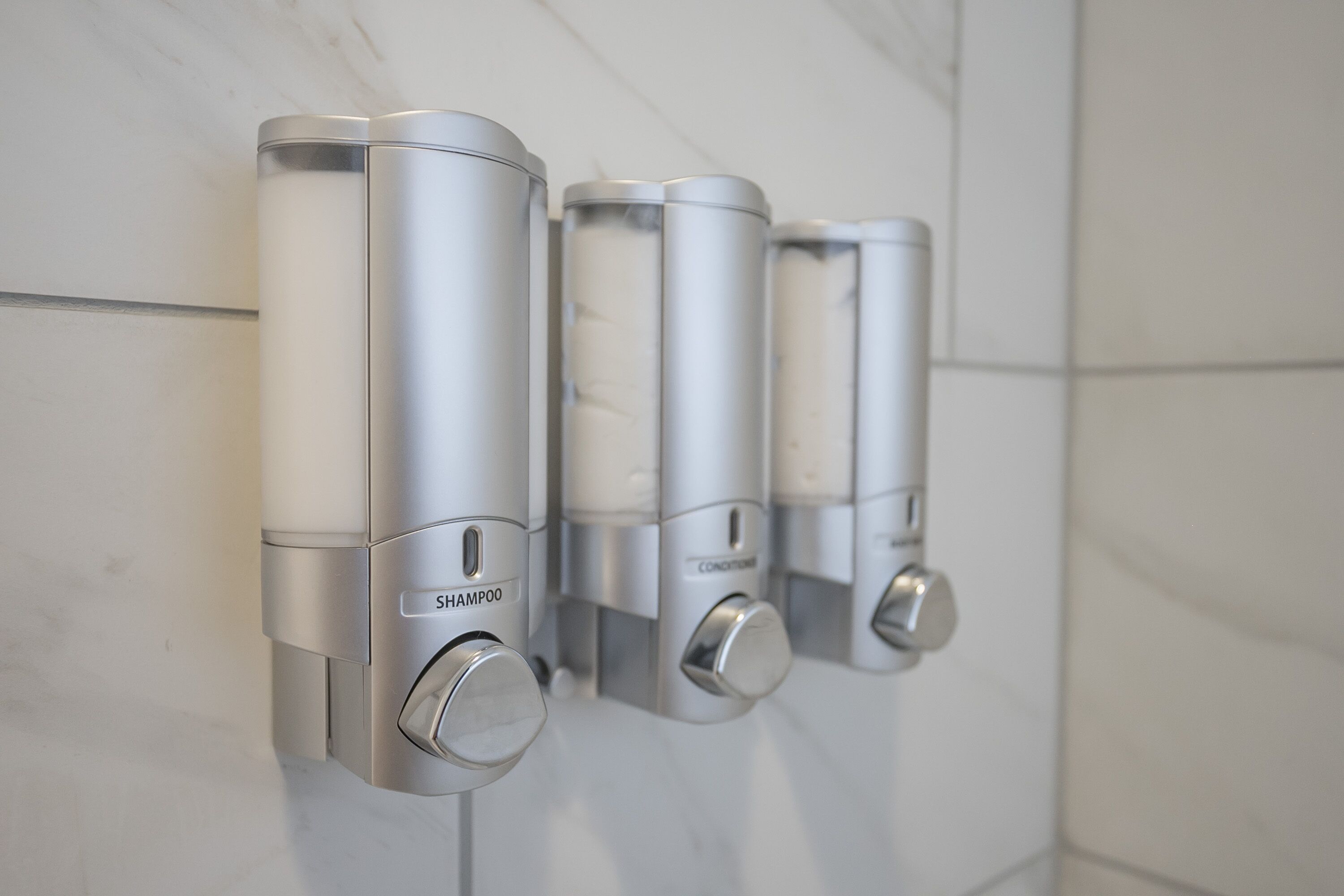 Three wall-mounted dispensers with a metallic finish, labeled for shampoo and conditioner.