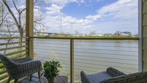A wooden deck with wicker chairs and a potted plant, overlooking a calm river with a bridge and buildings in the distance. - ❤️ of DWNTWN, Riverfront w pool, walk 2 Broadway (Nashville)