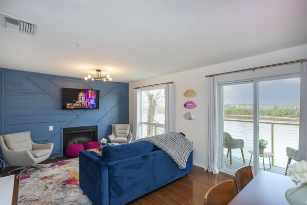 A modern living room with a blue sofa, a fireplace, a flat-screen TV, and a balcony with outdoor seating. - ❤️ of DWNTWN, Riverfront w pool, walk 2 Broadway (Nashville)