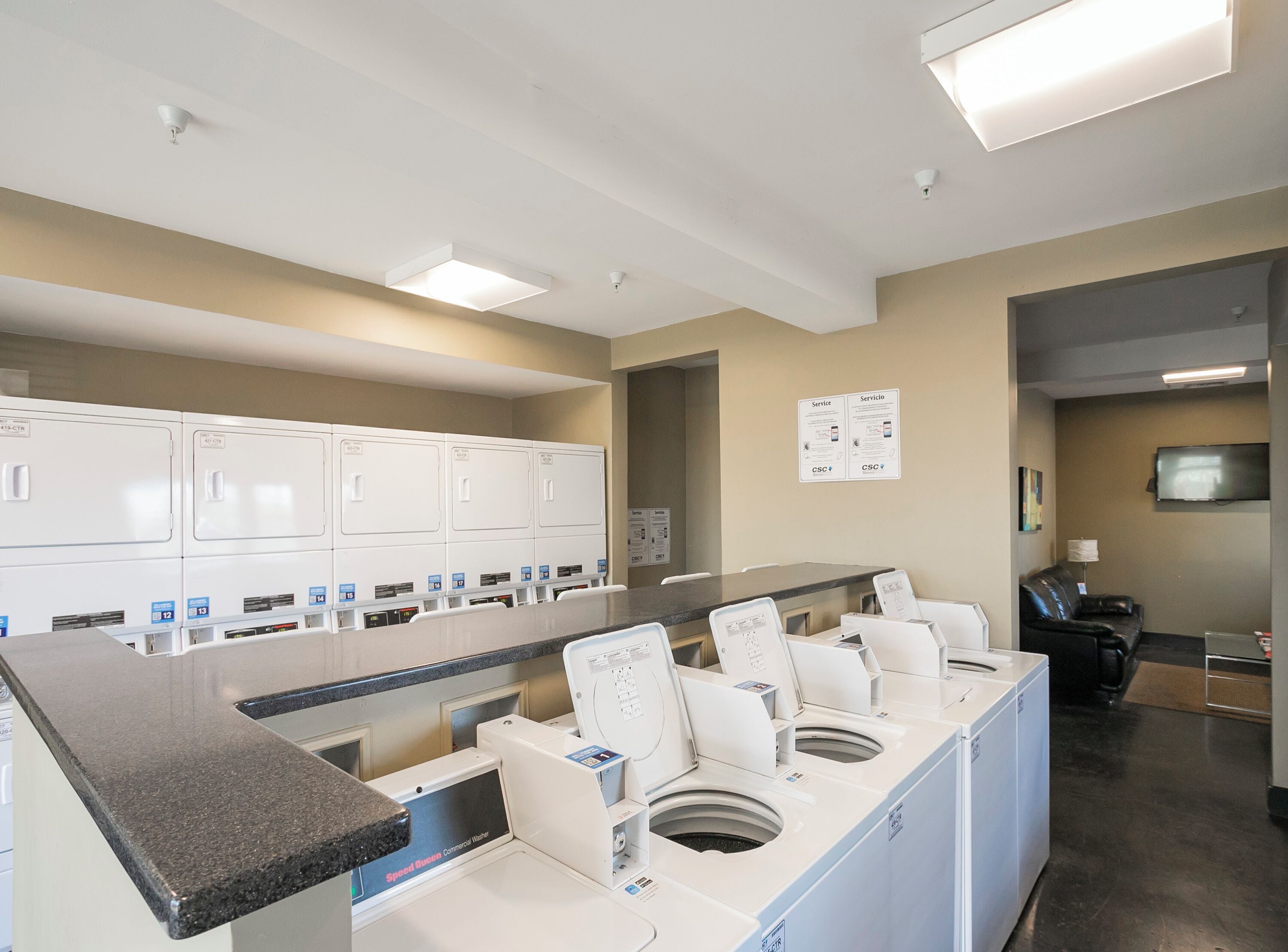 A laundry facility with multiple washing machines and dryers, a service information board, and a seating area with a television.