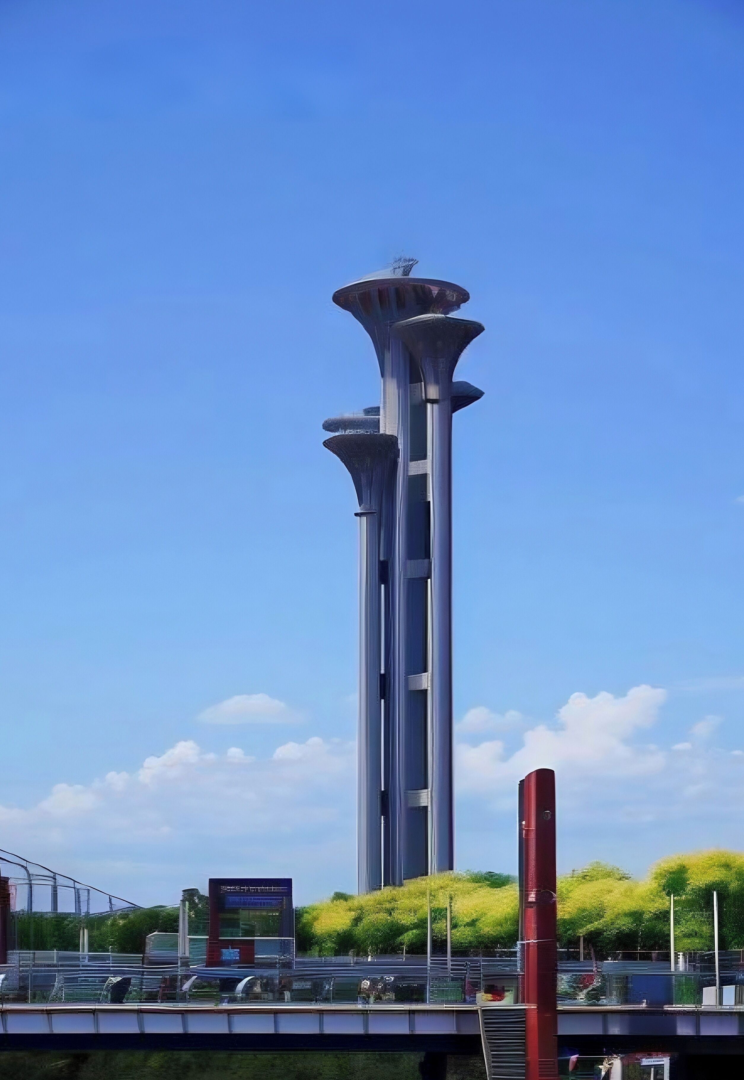 A tall, modern tower with a unique design, surrounded by greenery and a clear blue sky.