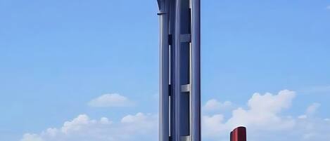 A tall, modern tower with a unique design, surrounded by greenery and a clear blue sky.
