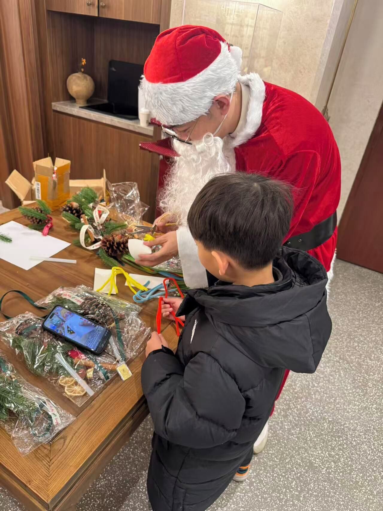 Une personne vêtue d’un costume de Père Noël est assise à une table, devant laquelle se trouvent divers objets, notamment un téléphone intelligent et des décorations.