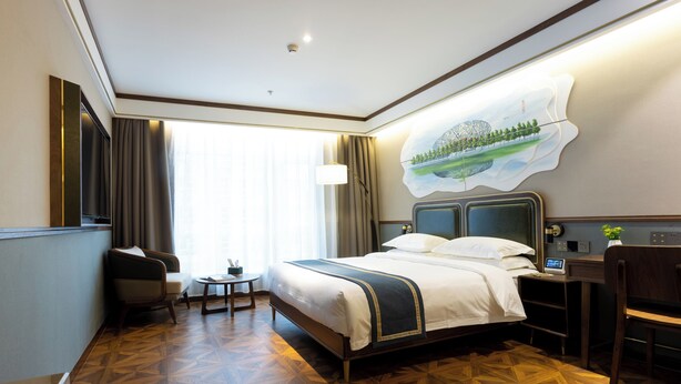 A hotel room with a large bed, a desk, a chair, a small table, and a painting on the wall.