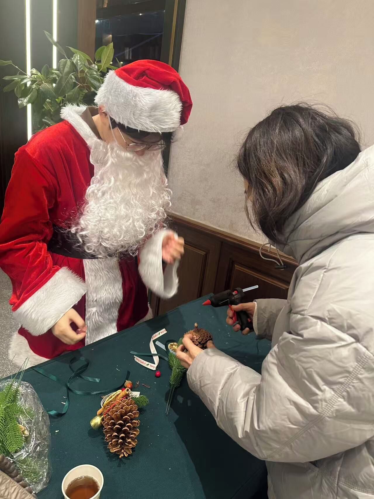 Una persona disfrazada de Papá Noel interactuando con otra persona en una mesa con mantel verde, piñas y una taza de té.