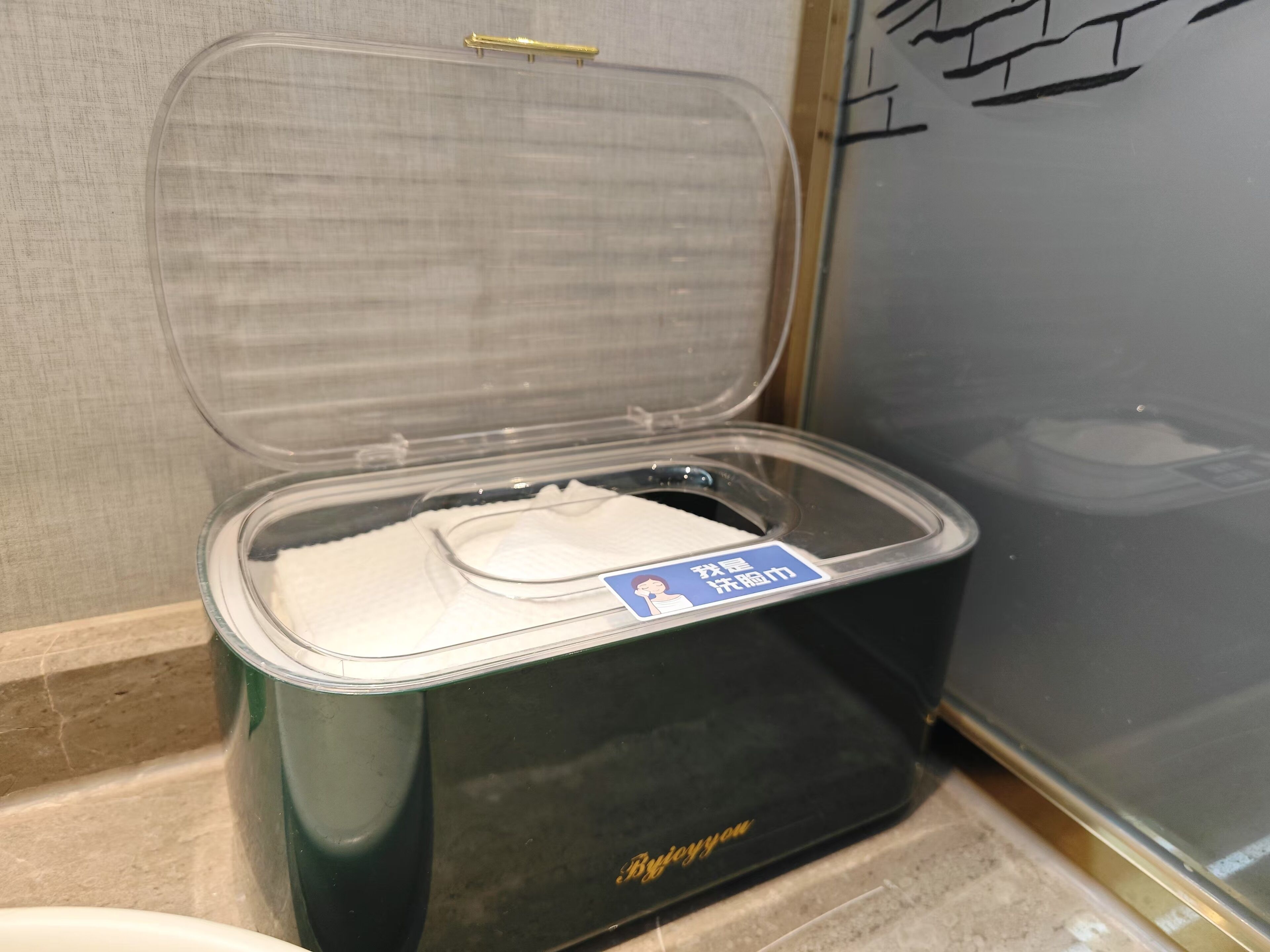 Un baño portátil negro con tapa transparente, un letrero azul con la figura de una mujer, y el logotipo "Byjoyyou".