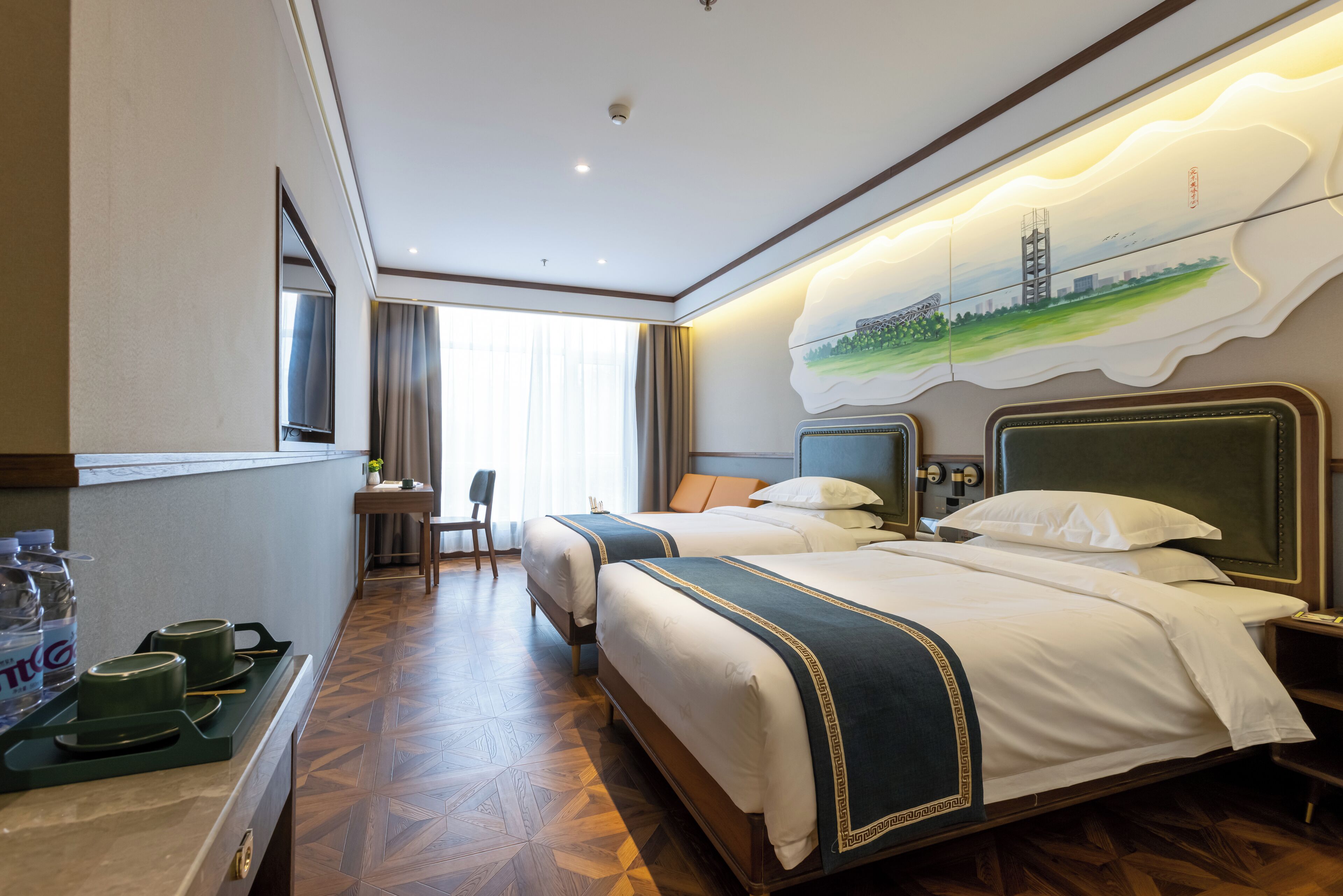 Ein Hotelzimmer mit zwei Betten, einem Schreibtisch und einem Stuhl. Es gibt ein Fenster mit Vorhängen, ein Bild an der Wand und eine Ablage mit Wasserflaschen und Gläsern.