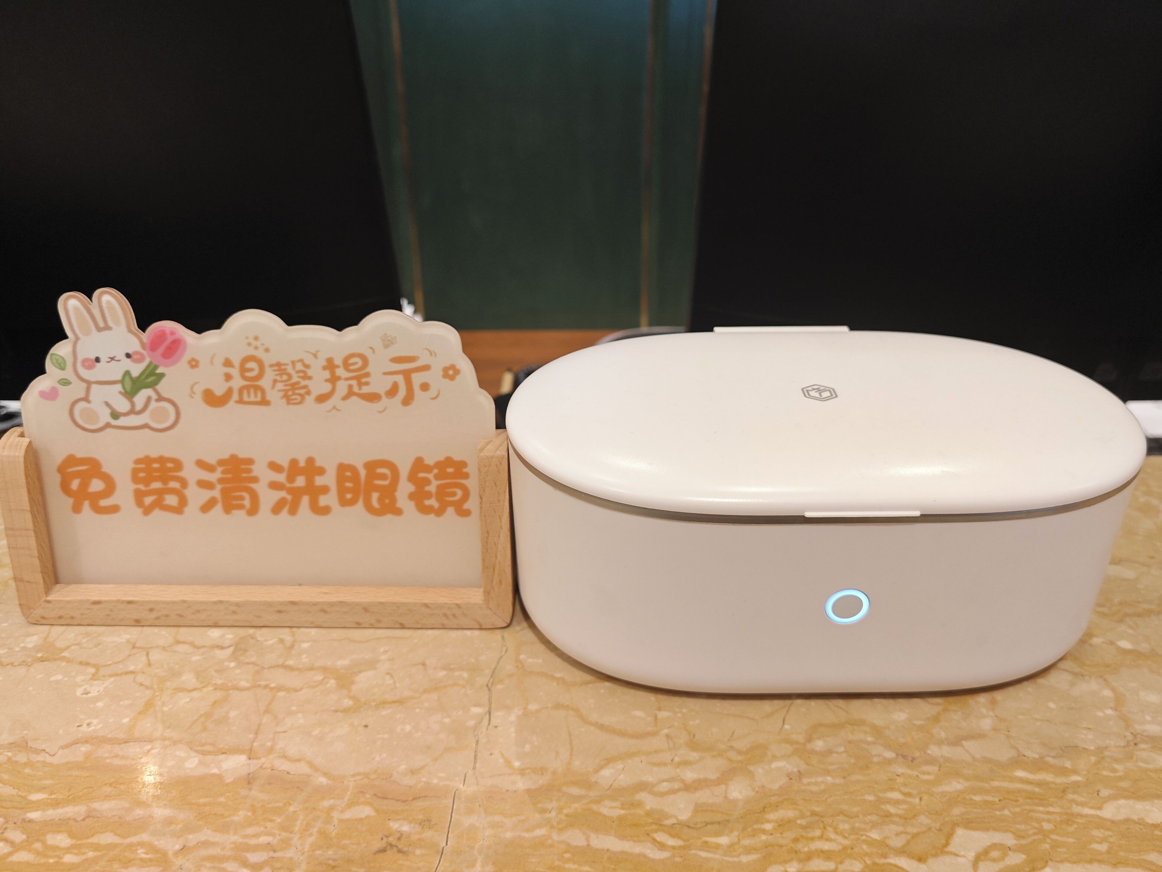 A white portable device with a blue light indicator, placed on a marble floor next to a wooden stand with a sign featuring a cartoon rabbit and Chinese text.