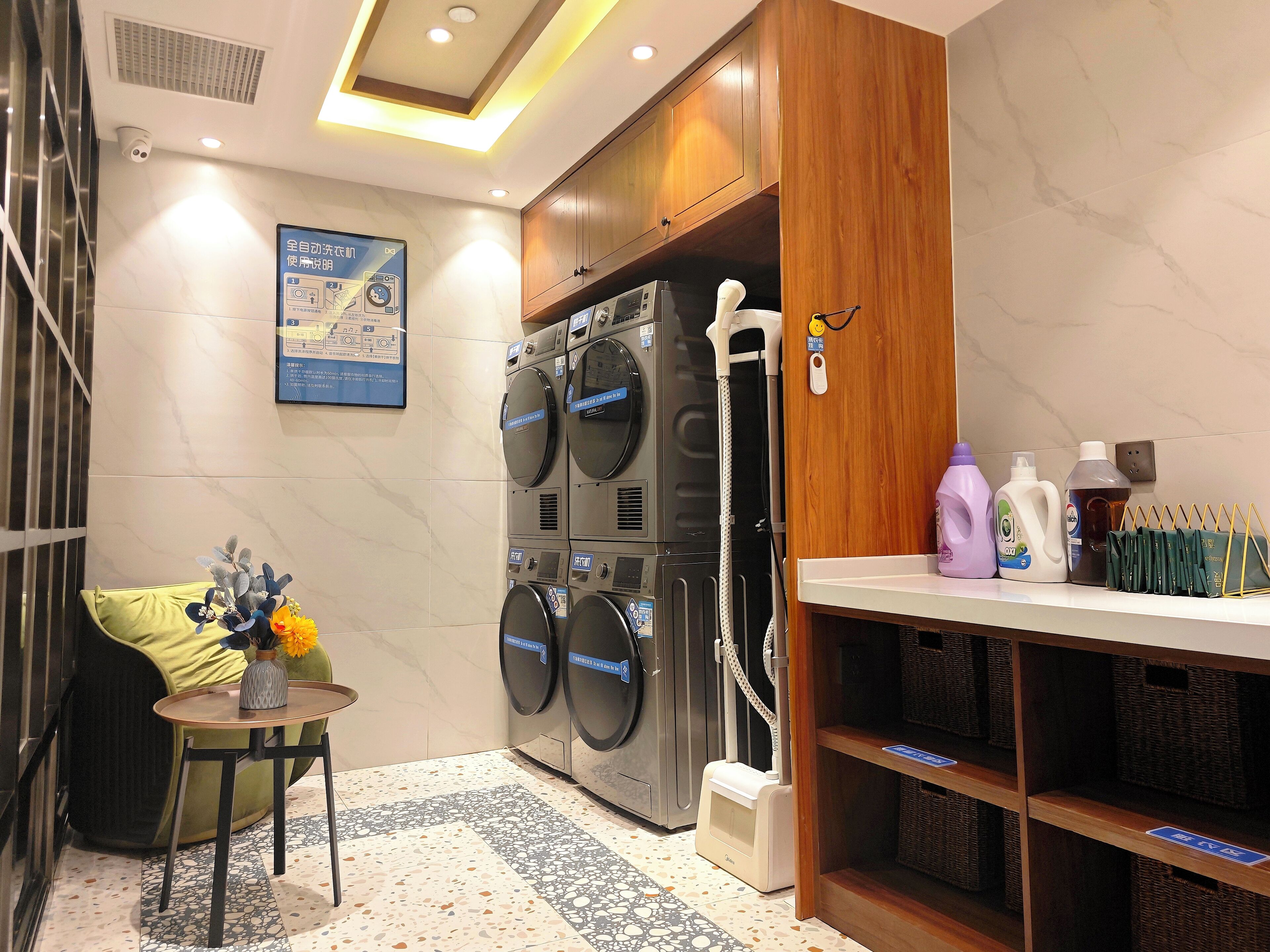 A laundry room with multiple washing machines, a table with a vase of flowers, and shelves with cleaning supplies.