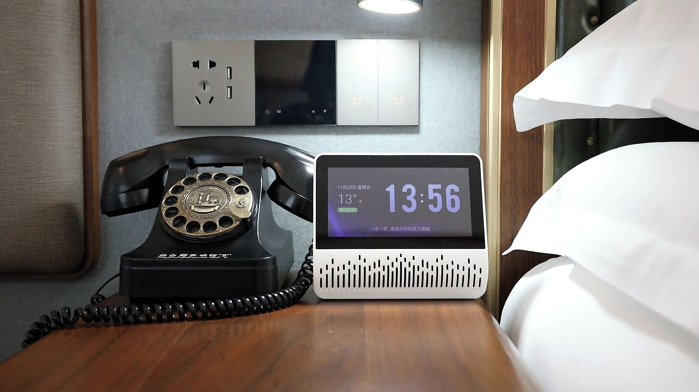 A hotel room with a rotary phone, a digital clock displaying the time and temperature, and a power outlet on the wall.