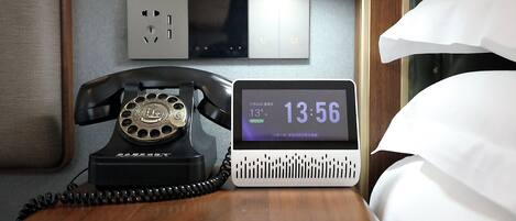 A hotel room with a rotary phone, a digital clock displaying the time and temperature, and a power outlet on the wall.