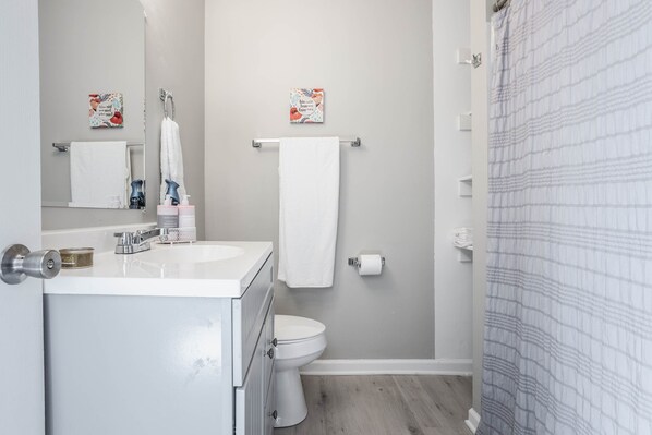A bathroom with a white sink, toilet, and shower curtain. There are two towels hanging on the wall and a small shelf with toiletries. - Grand Park, Westfield, Space For Entire Team, Sleeps 30 comfortably. Unit A (Sheridan)
