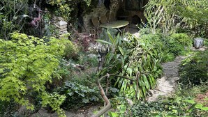 A garden with a stone pathway, lush greenery, and a rustic stone structure. - La Bergerie (MIRABEL AUX BARONNIES)
