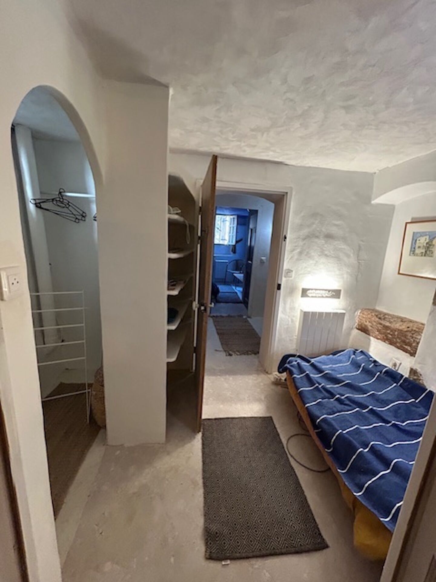 Ein schmales Zimmer mit einem Bett, einem Heizkörper und einer Tür zu einem anderen Raum.