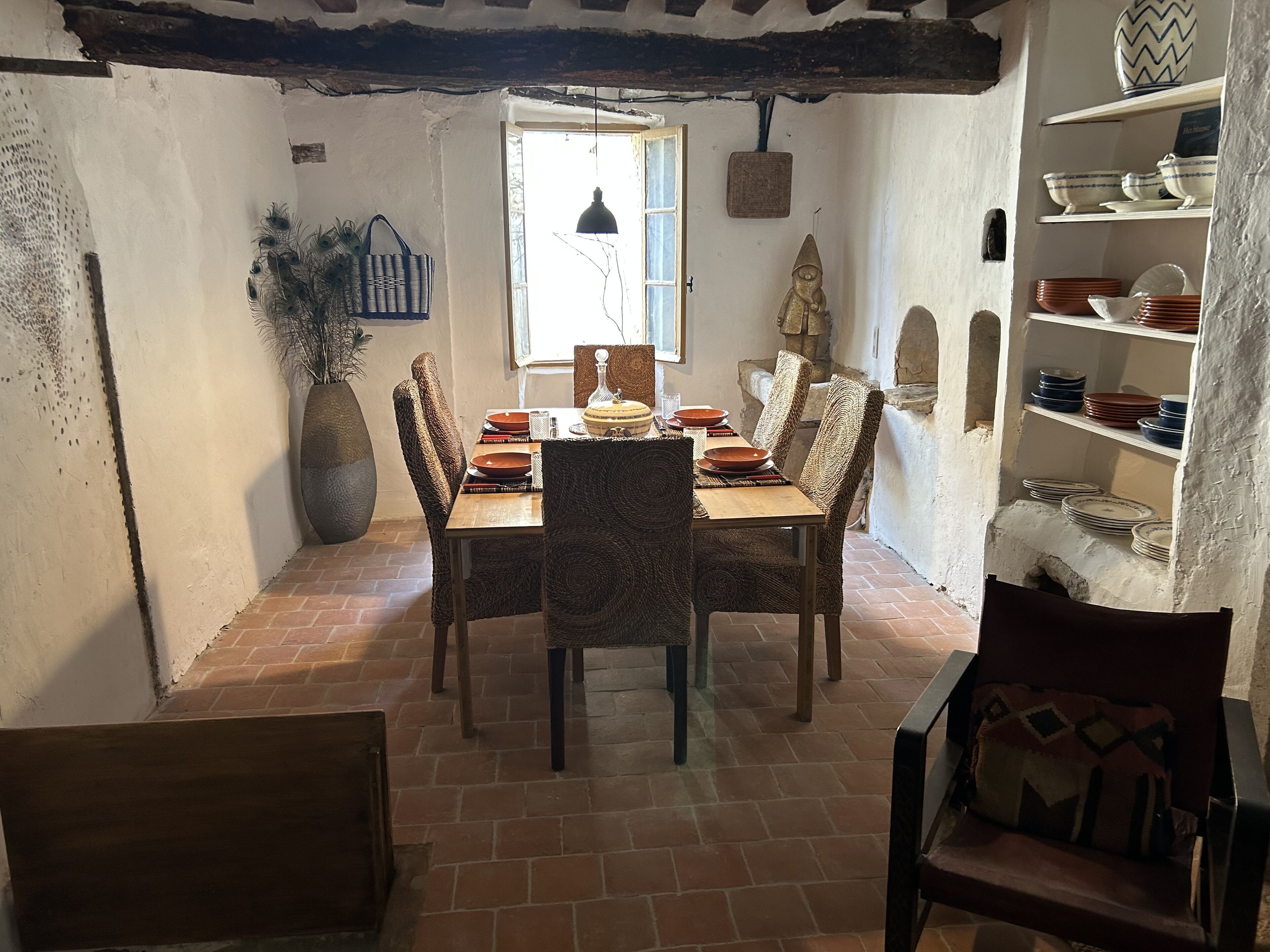 Un coin repas rustique avec une table en bois dressée pour quatre personnes, entourée de chaises en osier. Un grand vase et un panier sont posés sur la table, et une étagère remplie d’assiettes se trouve à droite.