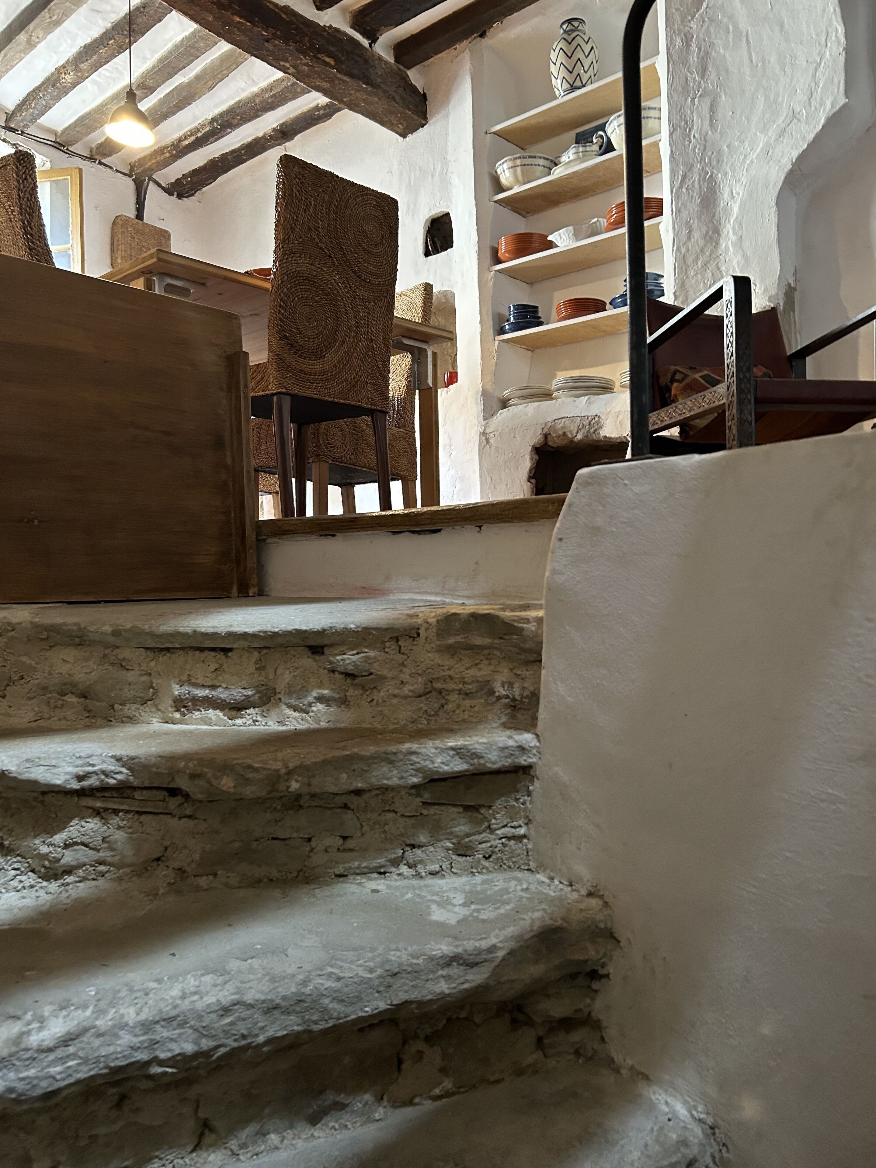 A rustic interior with stone steps leading to a wooden door, a woven chair, and shelves with dishes.