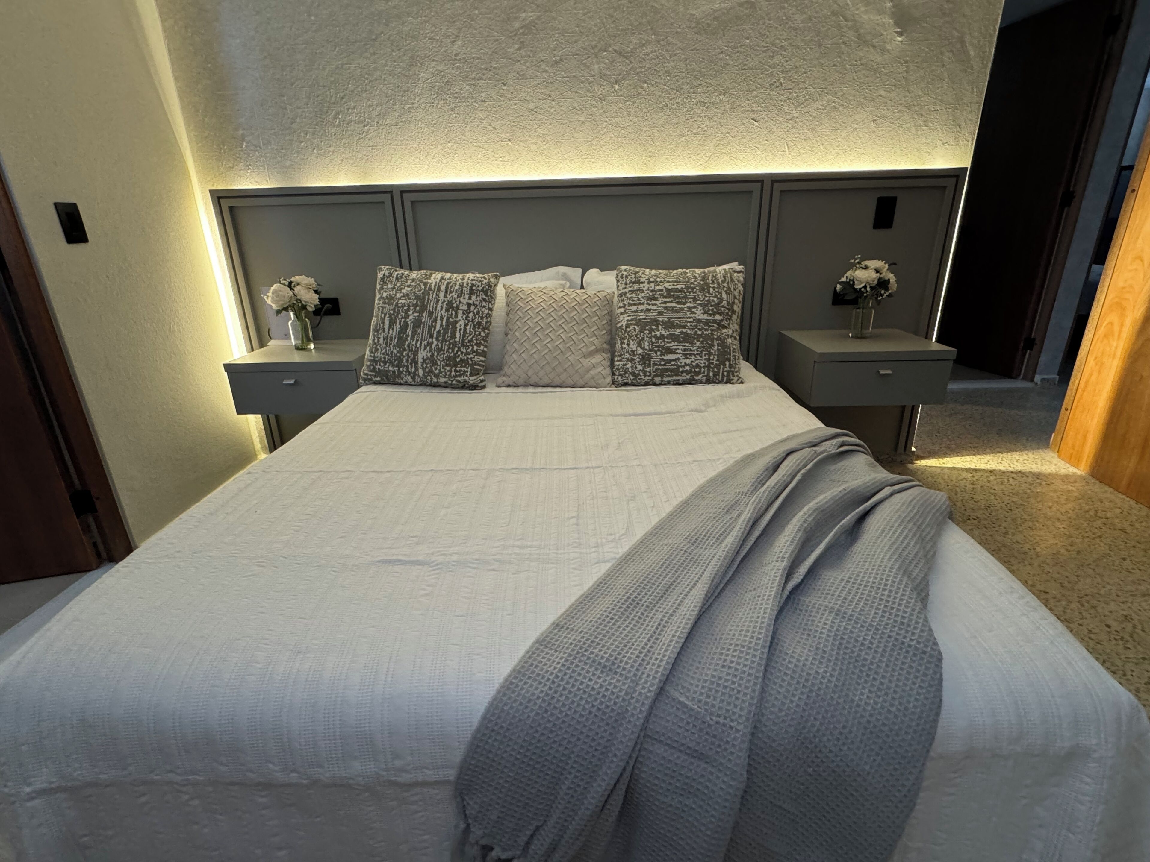 Un lit bien fait, recouvert d’une couette blanche et agrémenté de plusieurs oreillers, avec de chaque côté deux tables de chevet sur lesquelles sont posées des lampes et des compositions florales.