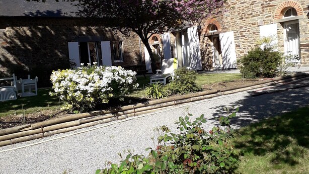 Un edificio de piedra con camino de grava, muebles de exterior blancos y plantas con flores.