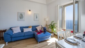Un salon moderne avec un canapé bleu, une table à manger dressée avec des assiettes et des verres, et une grande fenêtre offrant une vue sur la mer.