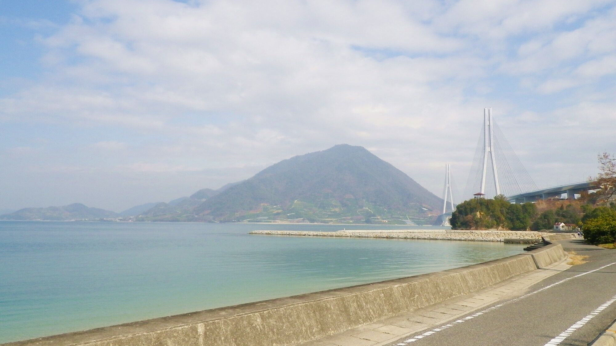 A coastal road with a bridge, a mountain, and a calm sea.