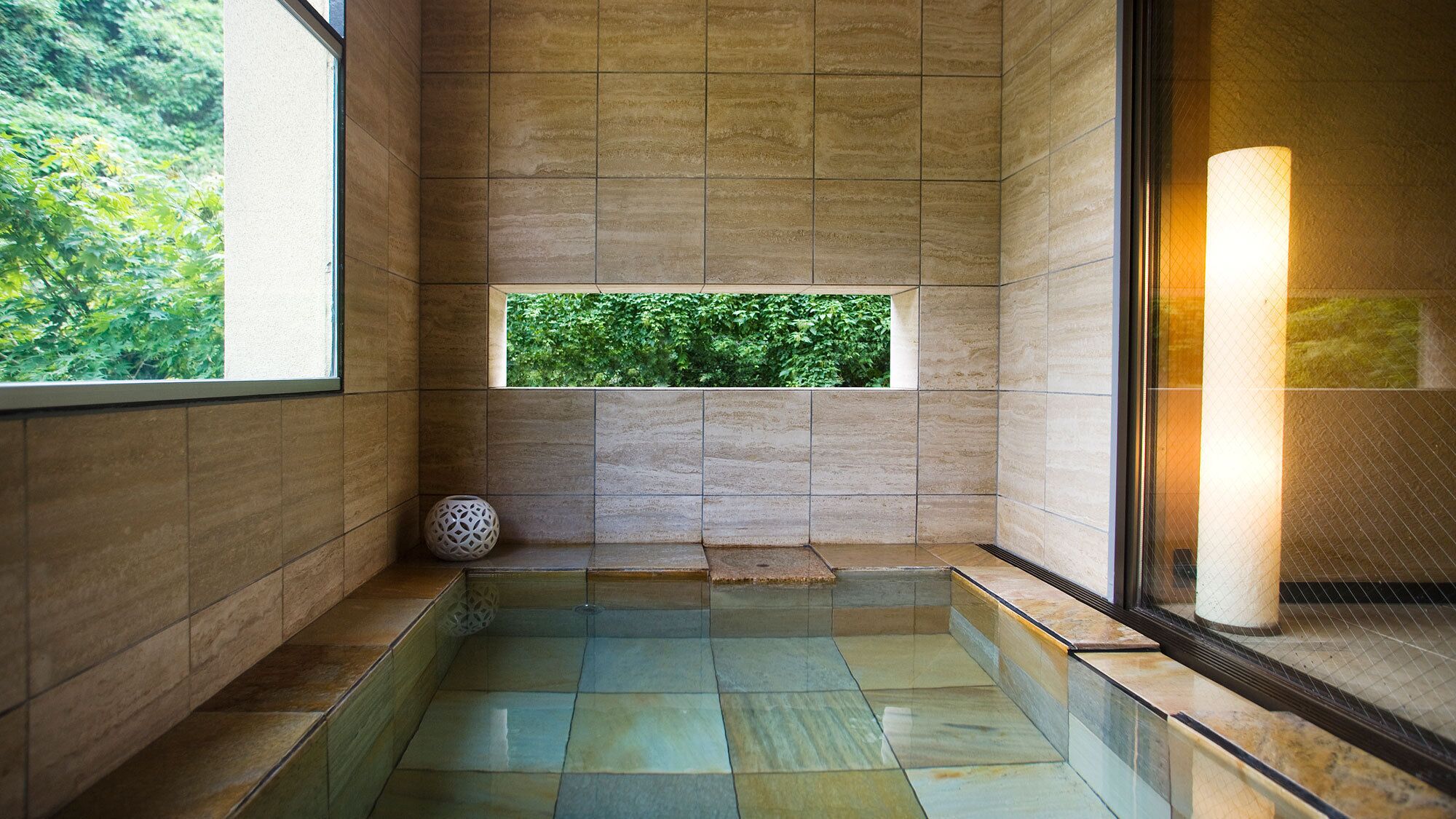 Ein modernes Badezimmer mit Glasboden, einem Fenster mit Blick auf Grünflächen und einer großen, zylindrischen Leuchte.