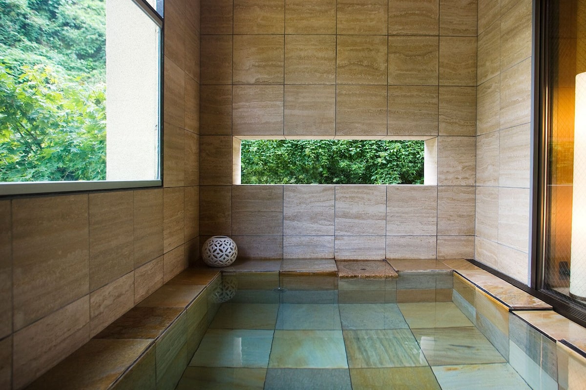 Un baño moderno con piso de vidrio, ventana con vista a vegetación y una lámpara cilíndrica grande.
