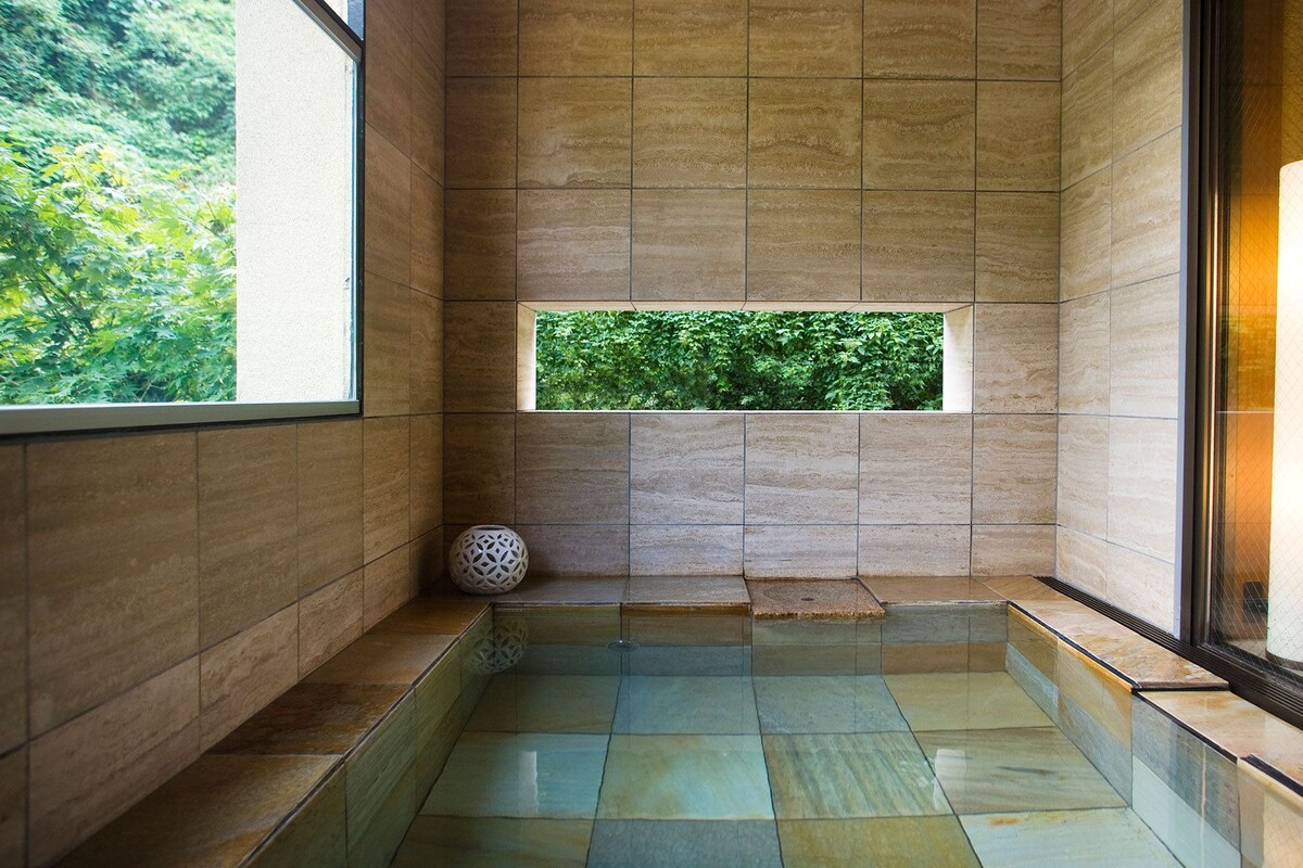 Un baño moderno con piso de vidrio, ventana con vista a vegetación y una lámpara cilíndrica grande.
