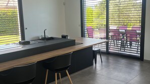 A modern kitchen with a large island, black bar stools, and large windows with blinds.