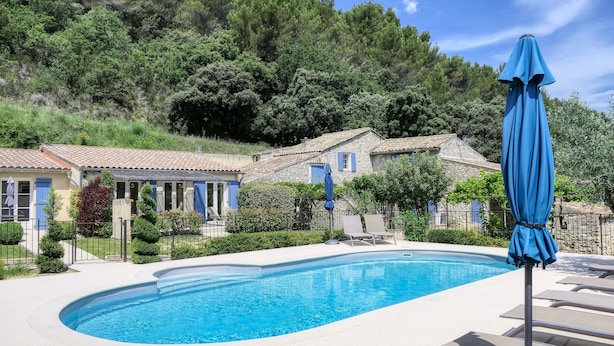 A swimming pool with lounge chairs and a blue umbrella, surrounded by stone buildings with blue shutters, set against a backdrop of lush greenery.