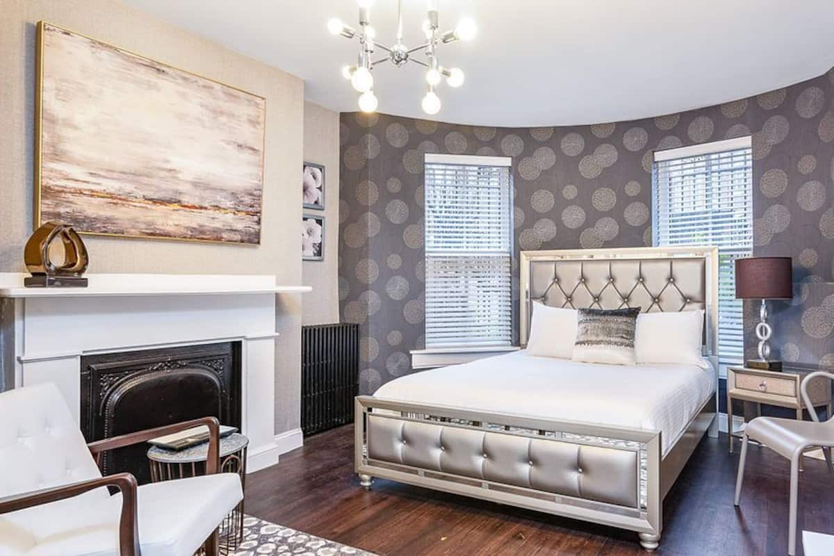 A bedroom with a large bed, a fireplace, two windows with blinds, a chandelier, and a side table with a lamp.