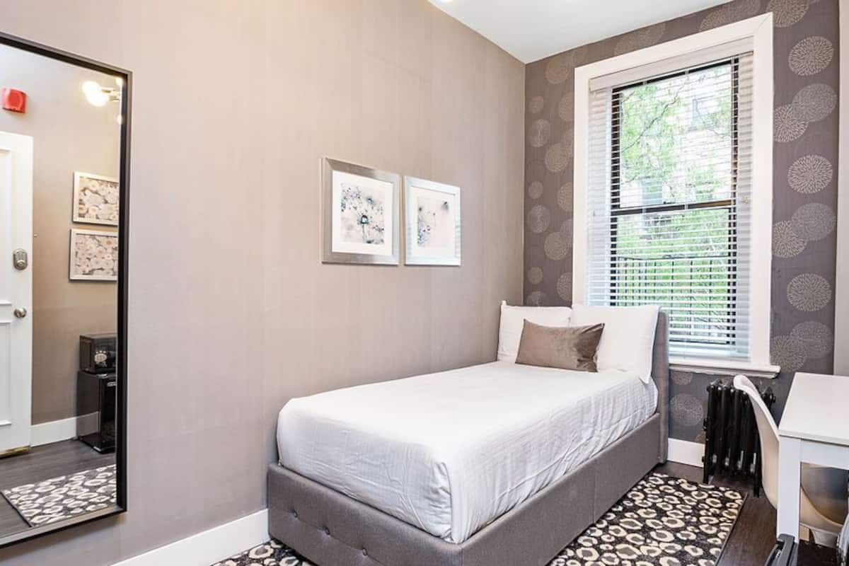A bedroom with a bed, a desk, a chair, a mirror, and a window with blinds.