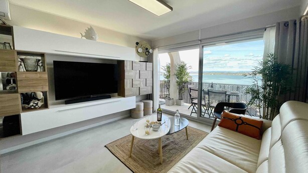A modern living room with a flat-screen TV, a sofa, a coffee table, and a balcony with a view of the sea.