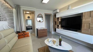 A modern living room with a sofa, a coffee table with glasses and bottles, a TV, and a kitchen area with a stove and oven.