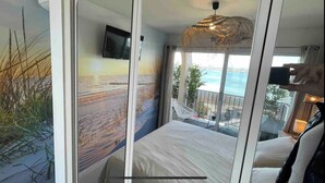 A bedroom with a large window, a bed, a TV, and a view of the sea.