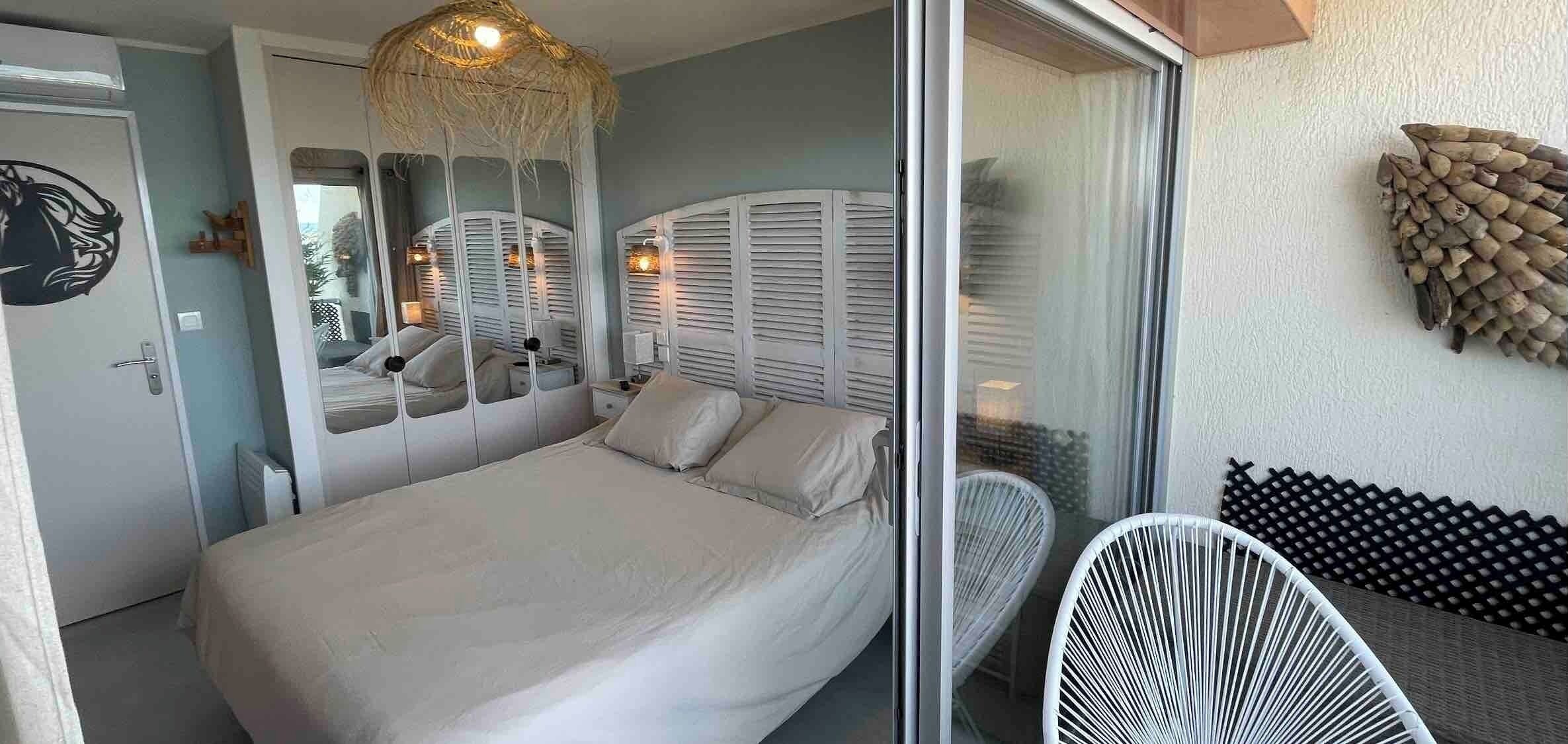 A bedroom with a bed, a large mirror, a hanging light fixture, and a view of the outside.