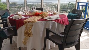 A dining table set for four with a red tablecloth and gold accents, chairs with cushions, and a view of the city through large windows. - Vigets Balou (Fort-de-France)